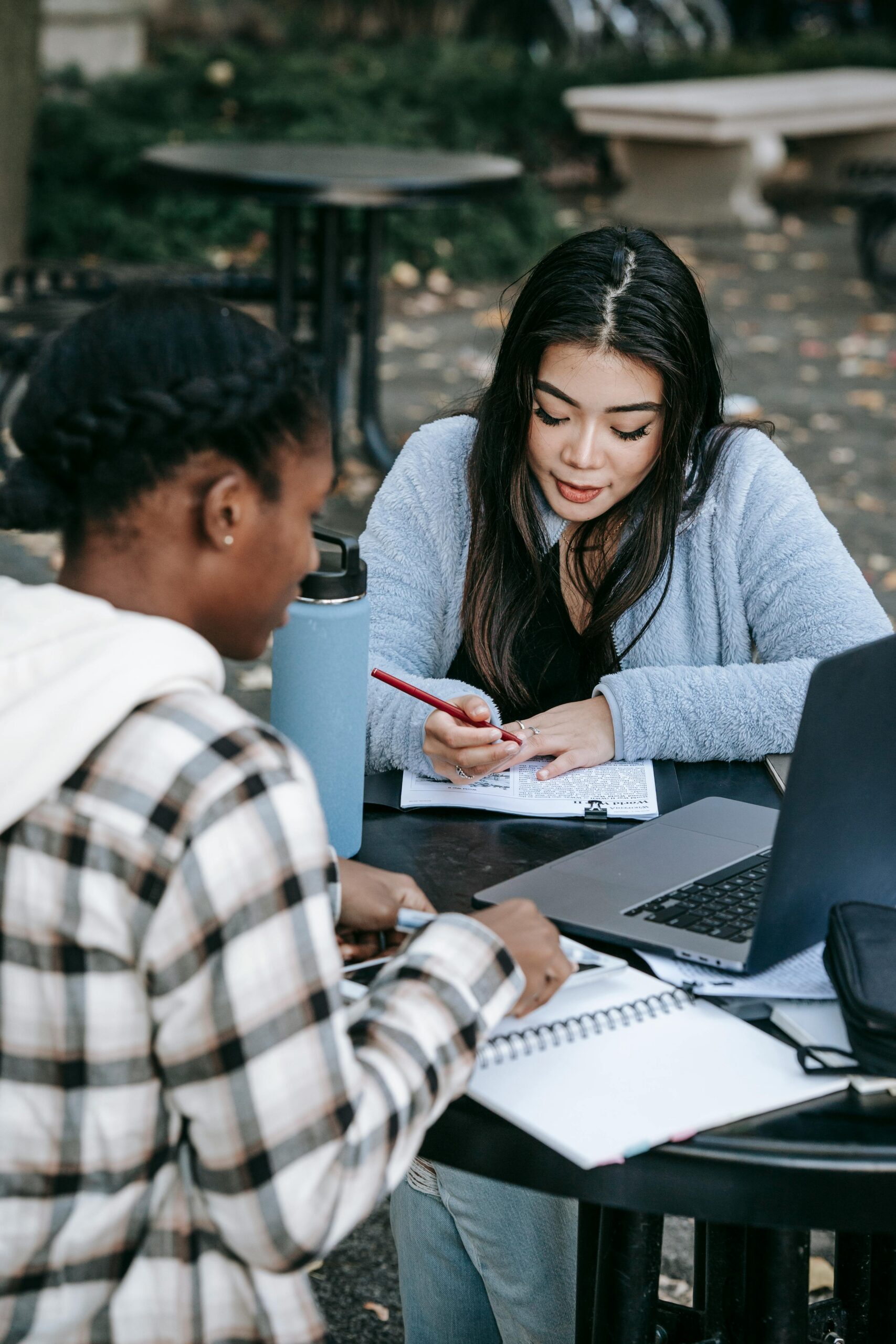 Two diverse students collaborating on a project outdoors with laptops and notes on a college campus.